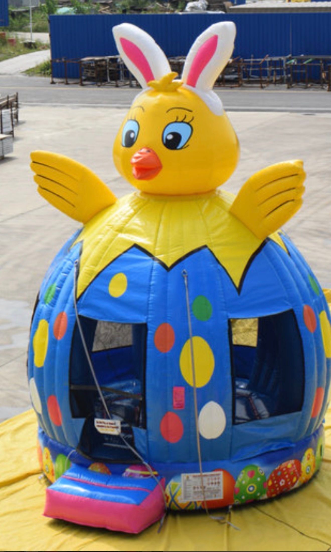 A blue, polka-dotted, inflatable Easter egg bounce house topped with a yellow chick wearing rabbit ears.