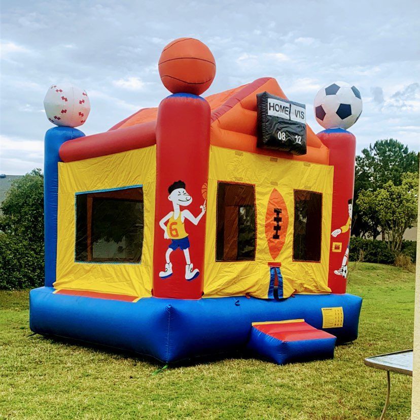 A colorful sports-themed inflatable bounce house with basketball, baseball, and soccer ball accents, set on a green lawn.