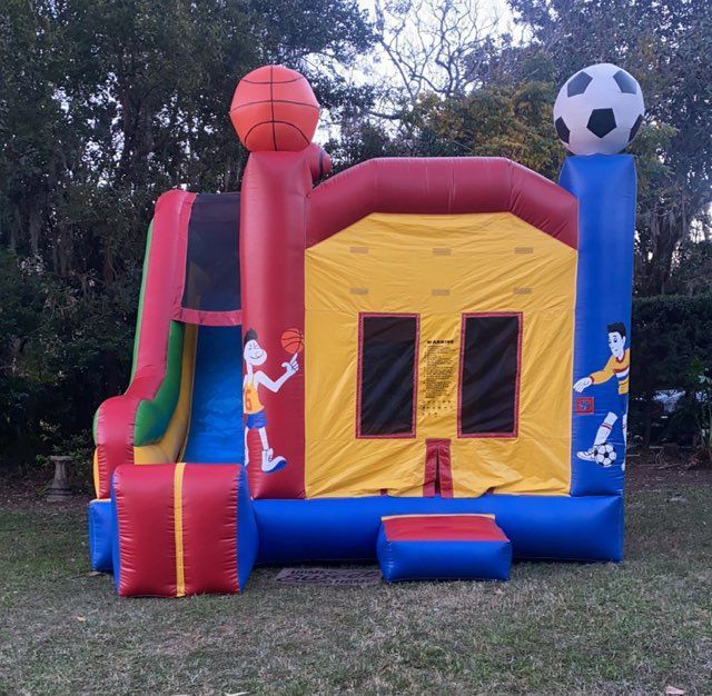 A red, yellow, and blue sports-themed inflatable bounce house with a slide, basketball, and soccer ball decor outdoors.