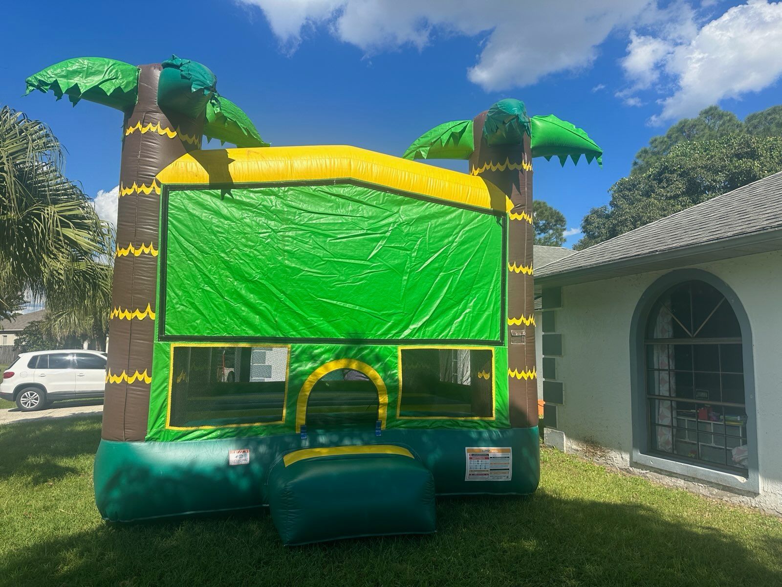 A green and yellow inflatable bounce house with palm tree pillars on a grassy lawn next to a house.