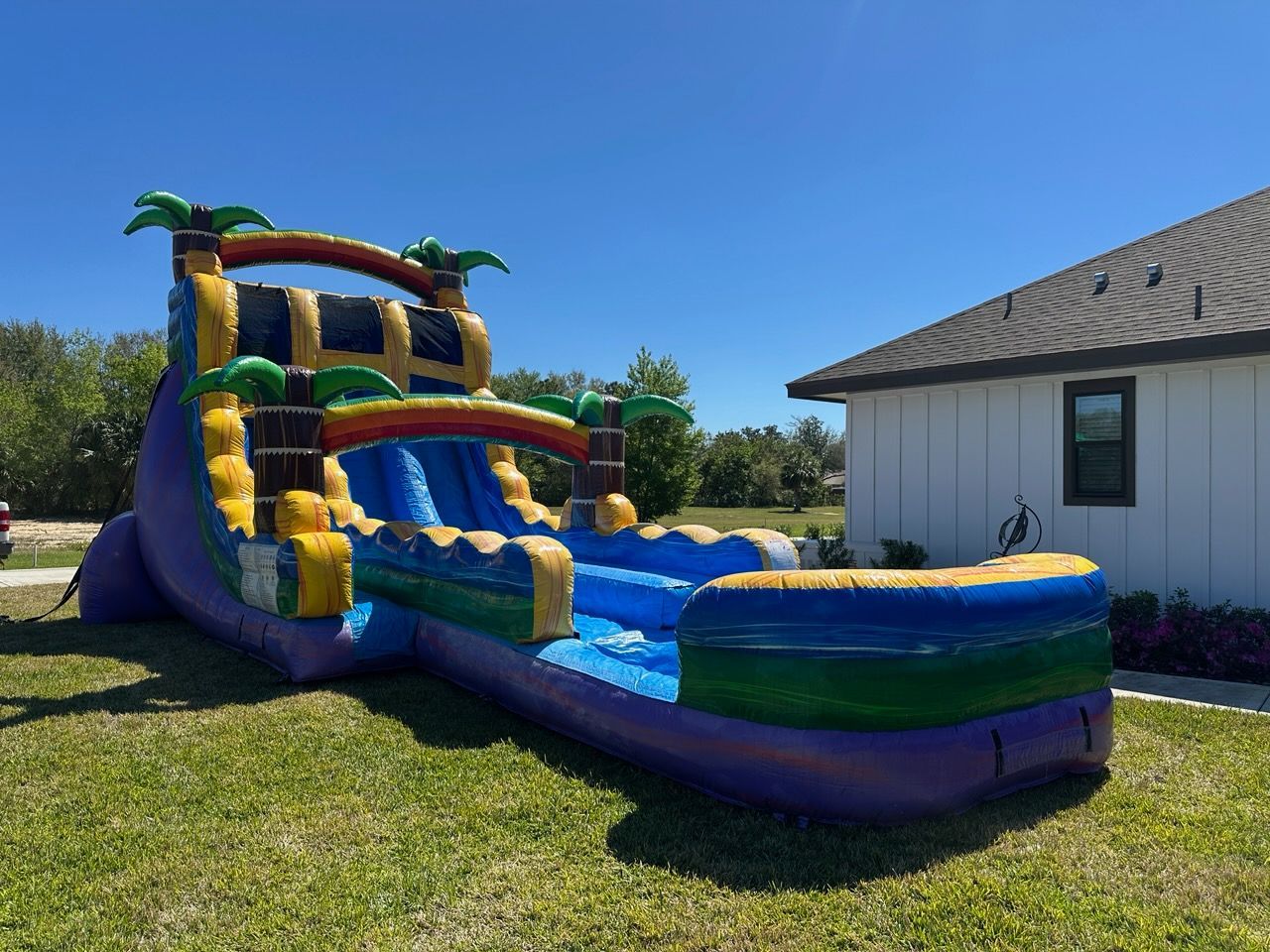 A colorful inflatable tropical-themed water slide set up on a grassy lawn next to a white house under a clear blue sky.