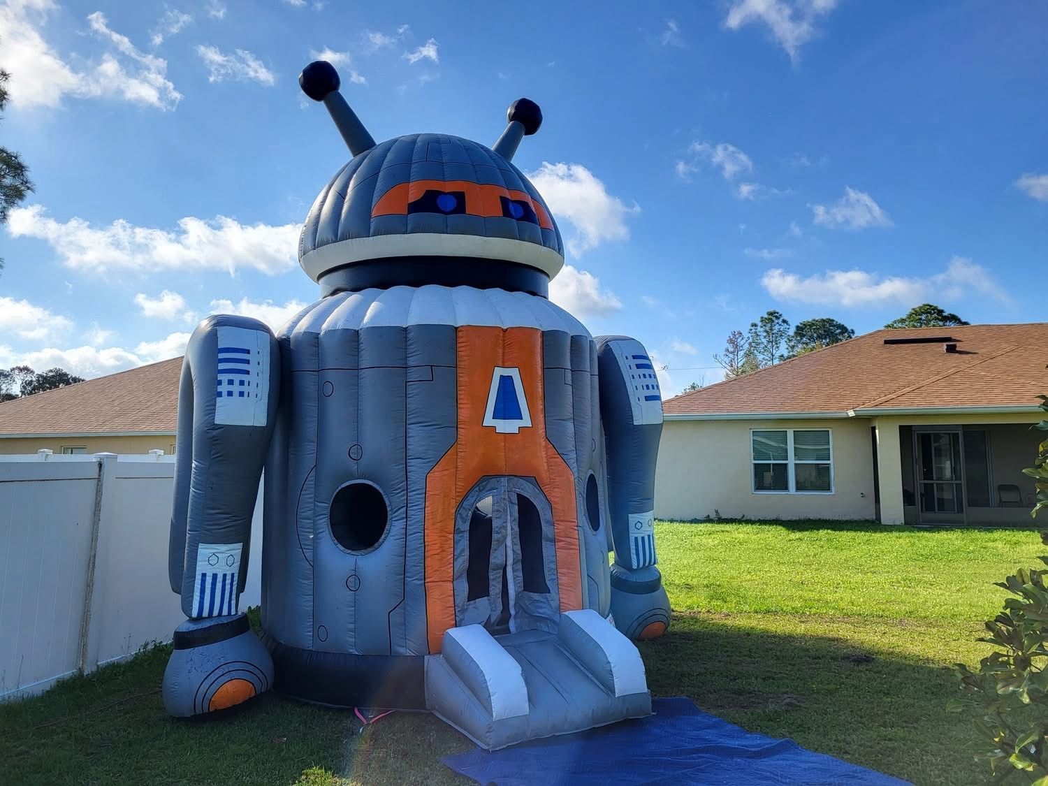 A large, inflatable robot-shaped bounce house in a backyard, featuring grey and orange panels with antennas.