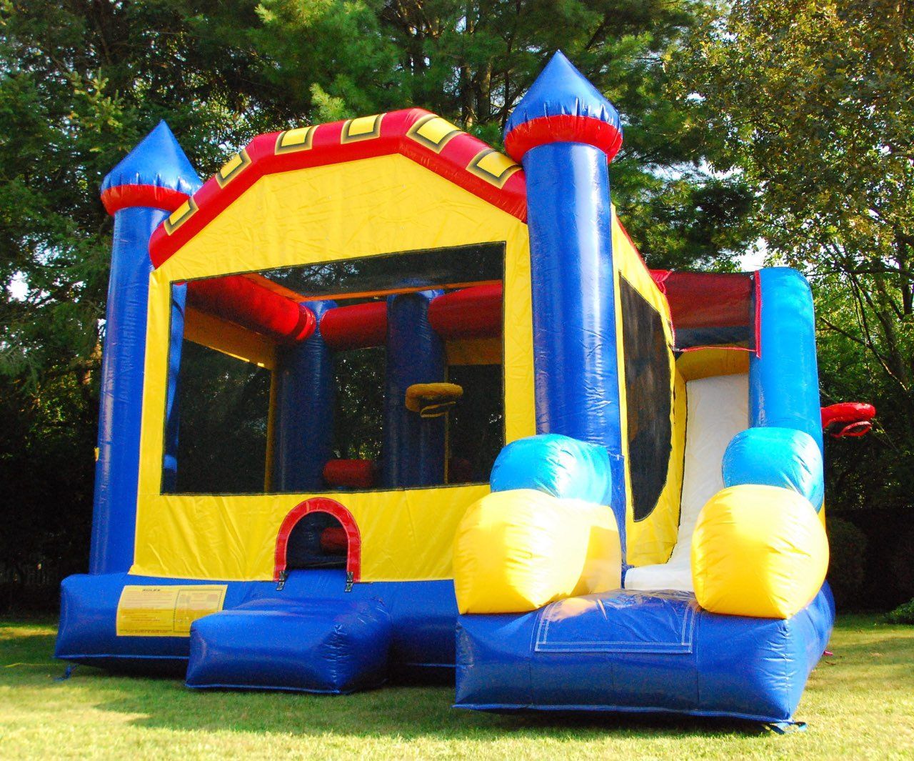 A colorful inflatable bounce house with a slide and basketball hoop, set on a grass lawn with trees in the background.
