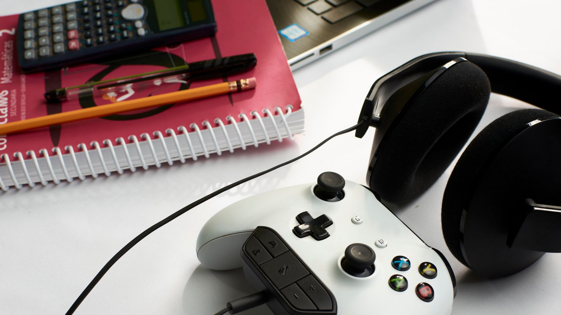 White desk with a white Xbox controller, headphones, a notebook with a calculator, and a laptop. White desk with a white Xbox controller, headphones, a notebook with a calculator, and a laptop.