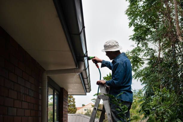 A man is standing on a ladder spraying a gutter with a hose.