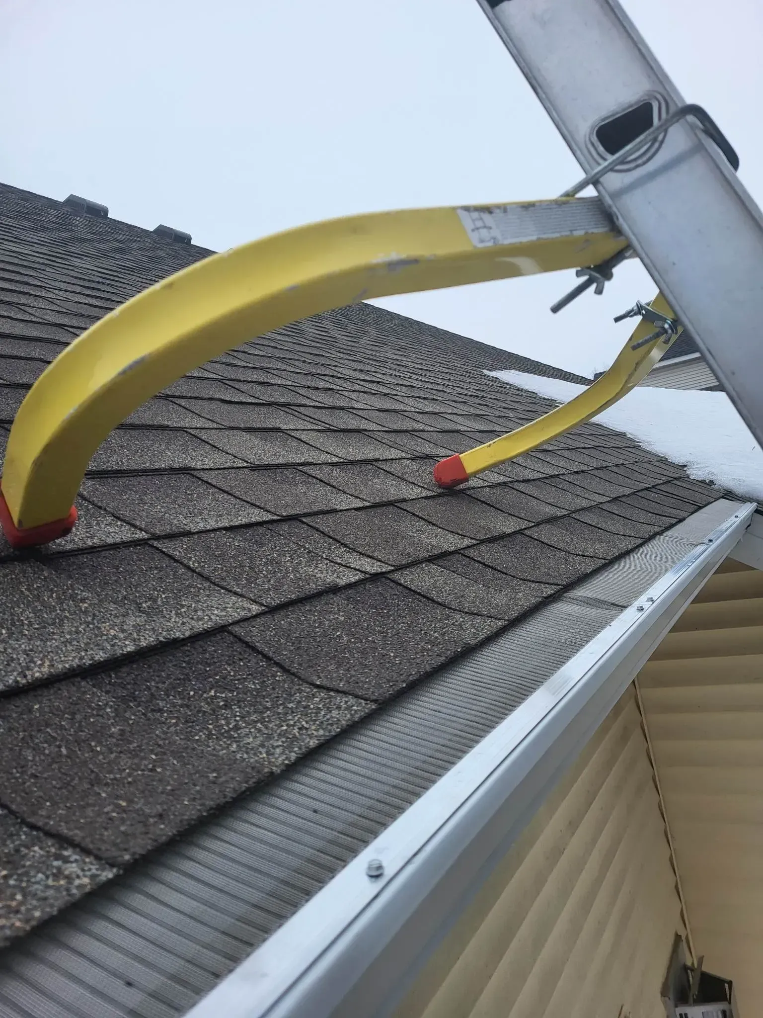 Yellow boom lift bucket resting against a gray shingle roof near the gutter under a cloudy sky