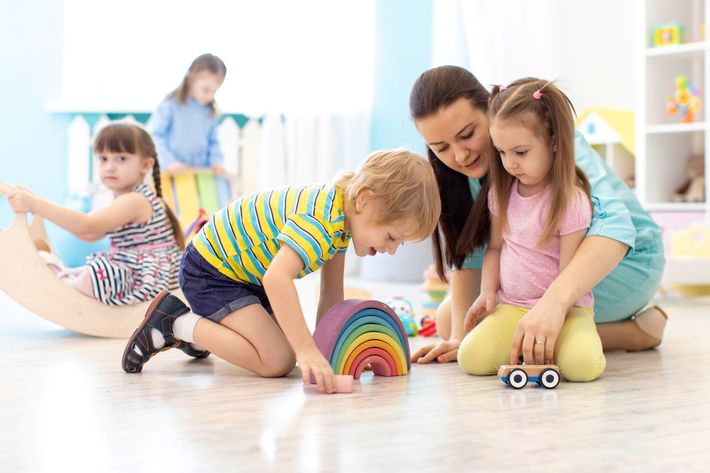 Children and adult playing with toys in a brightly lit room. A wooden rainbow, car, and arch are on the floor.
