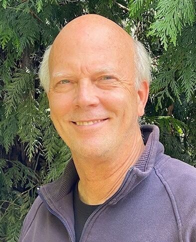 A bald man wearing a purple jacket is smiling in front of a tree.