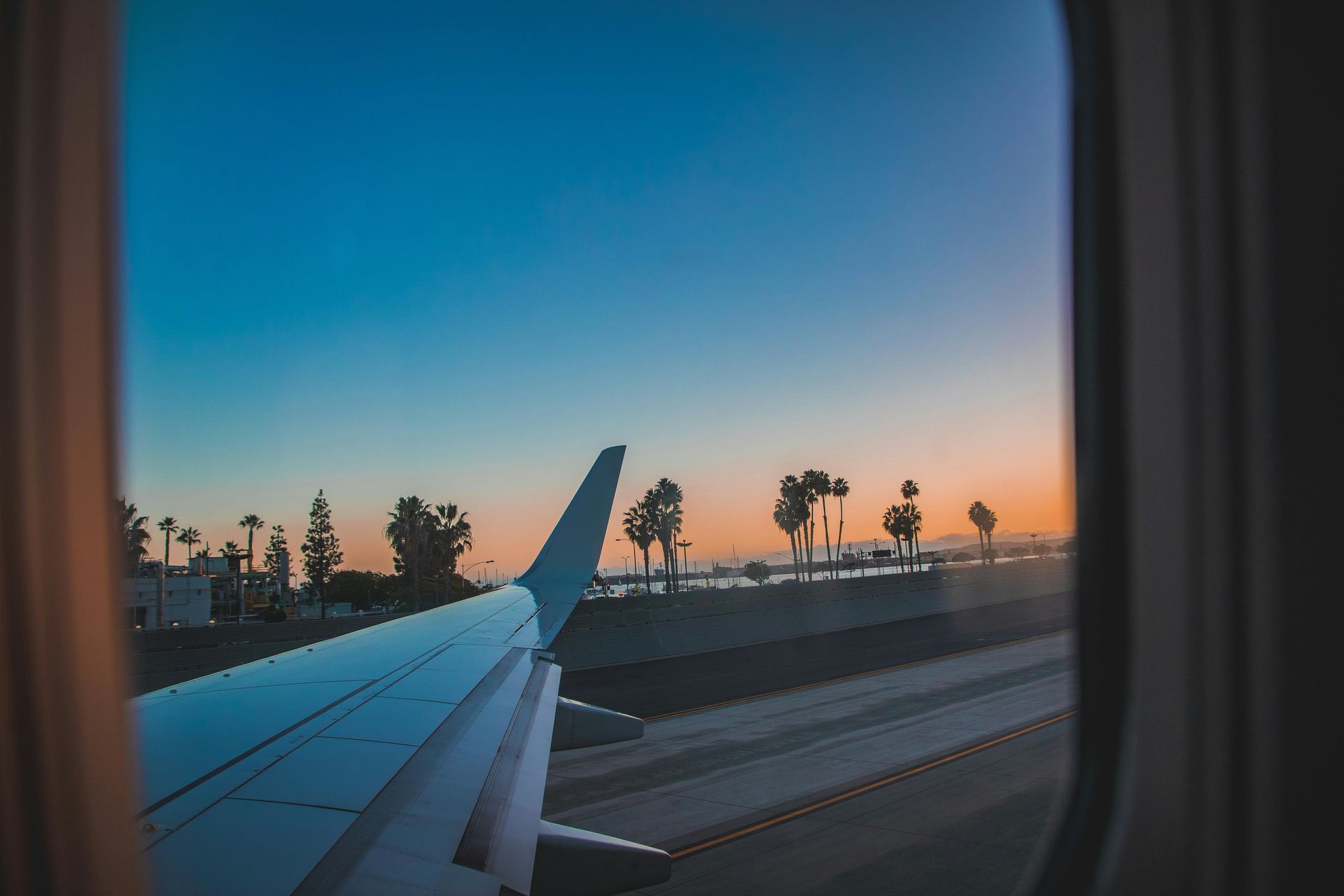 View from airplane window: wing against a colorful sunset sky; palm trees, runway visible.