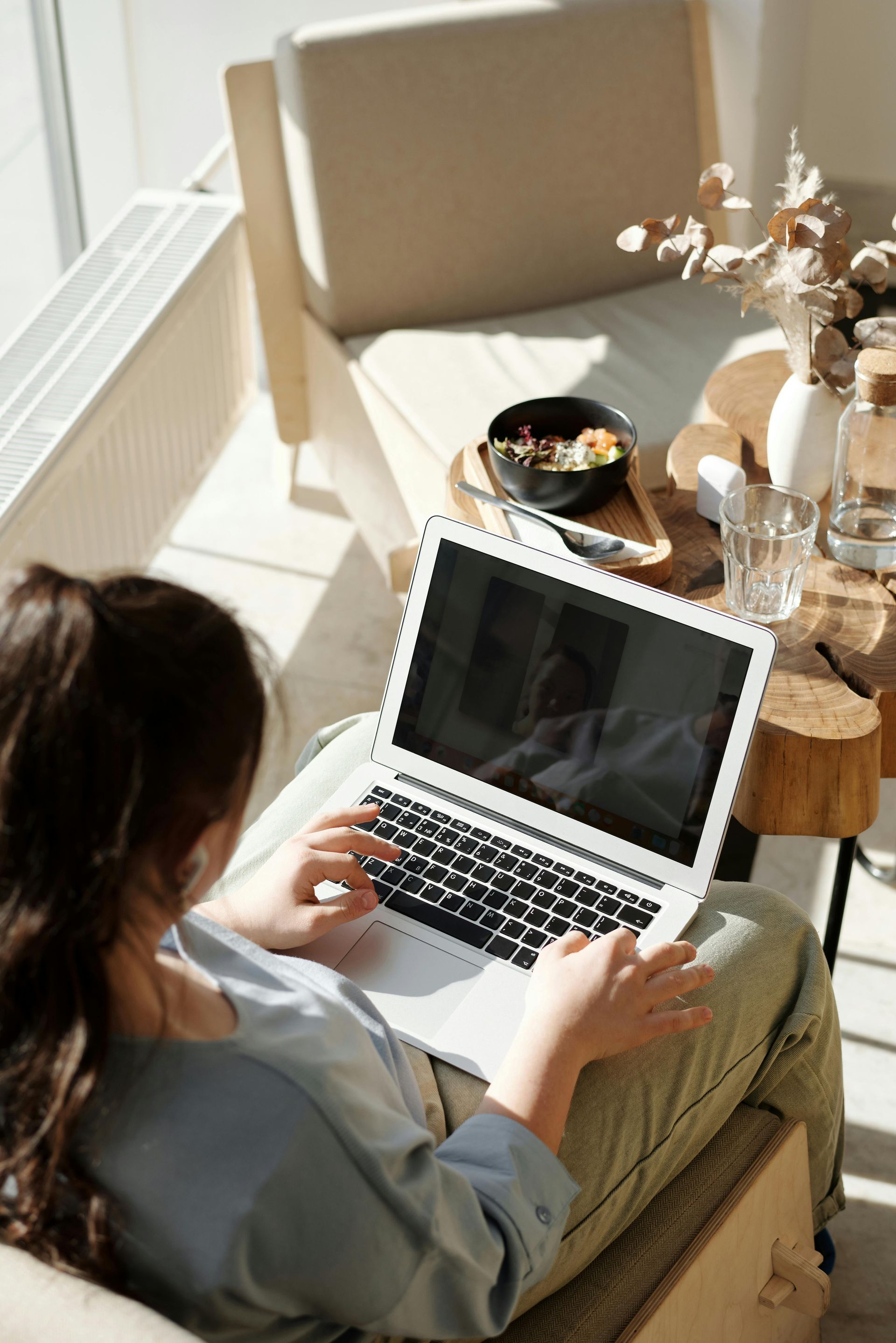 Woman typing on a laptop, seated on a chair near a window. Sunlight illuminates the scene.