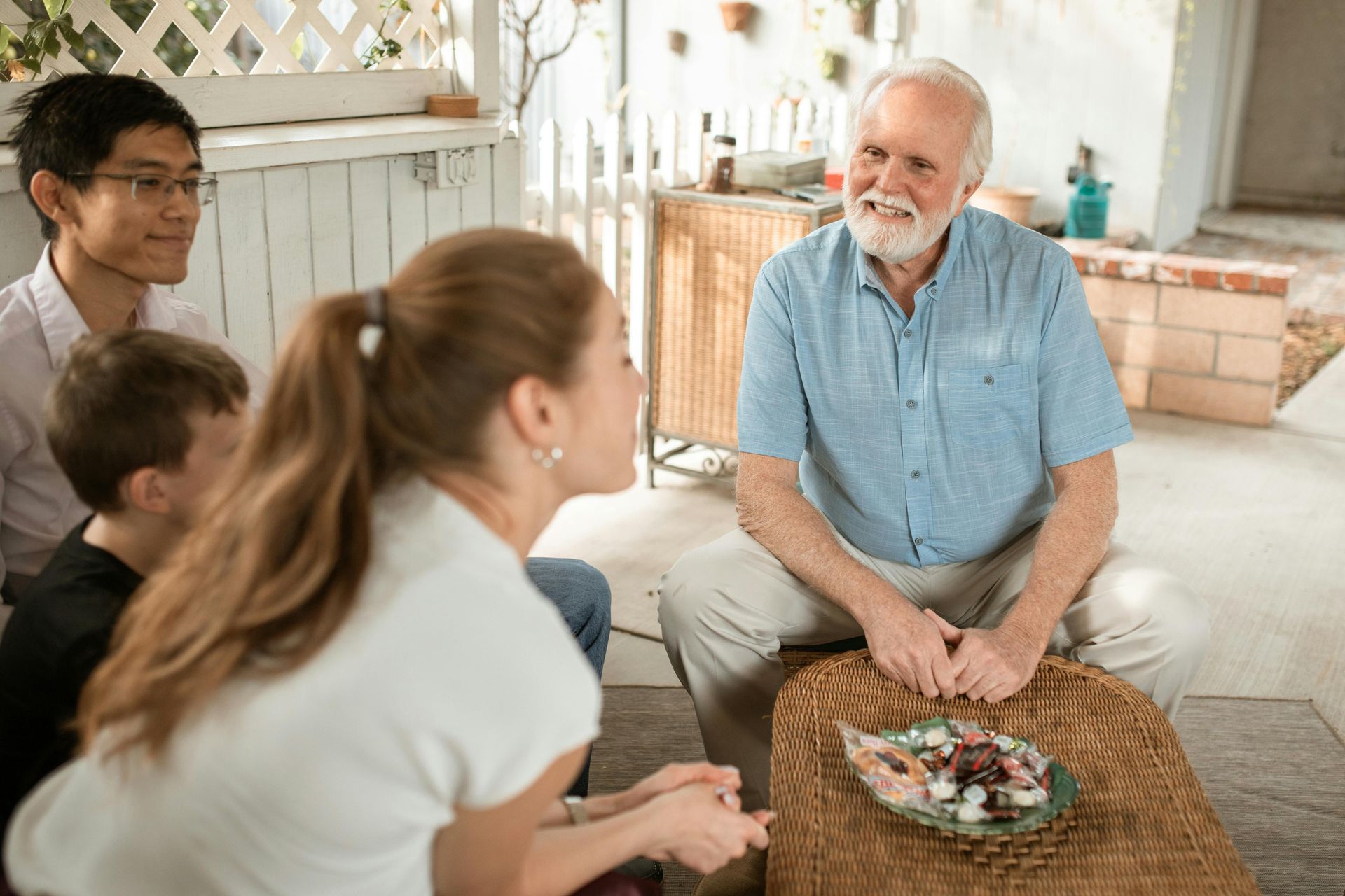 A group sits around a table outdoors. An older man smiles while looking at a plate. Others are engaged, attentive.