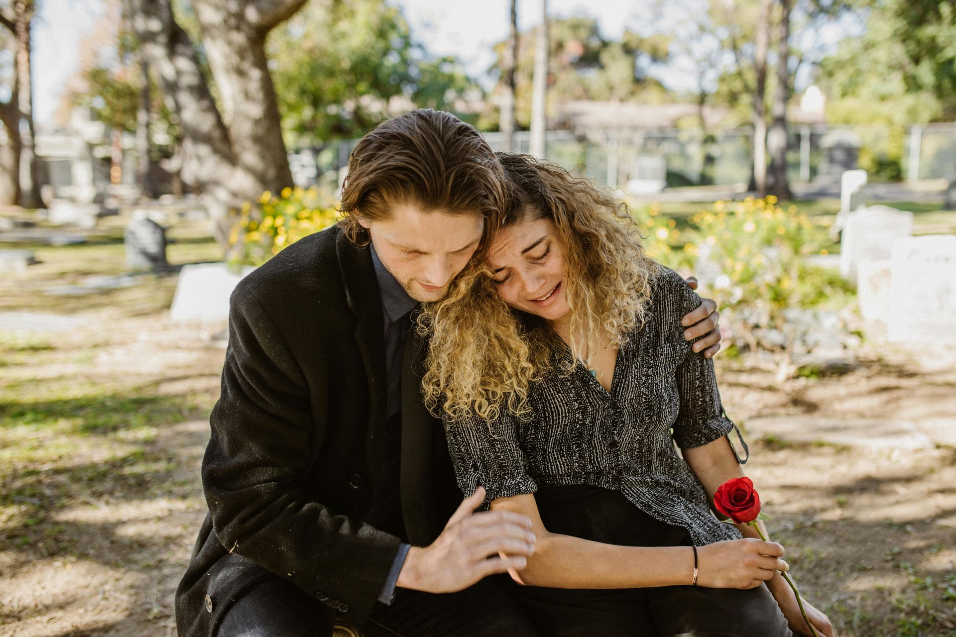 Man consoles a woman at a cemetery. She cries, holding a red rose.