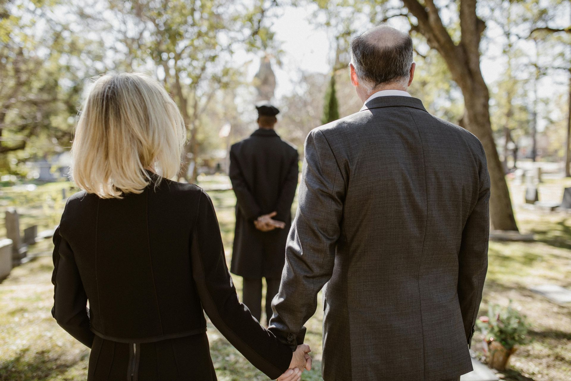 Couple holding hands, walking in a cemetery towards a person in a black coat.