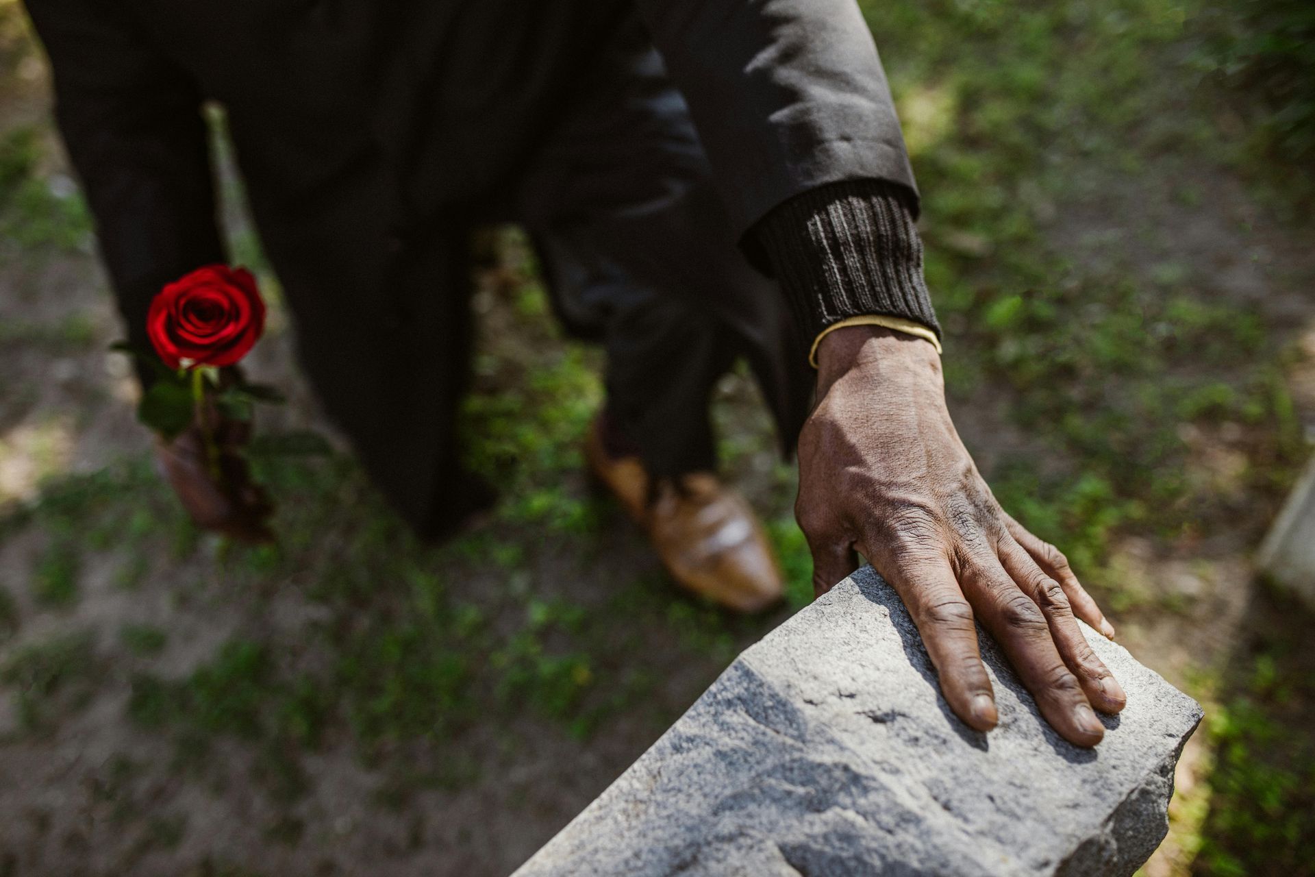 Person's hand on a headstone, holding a red rose at a cemetery.