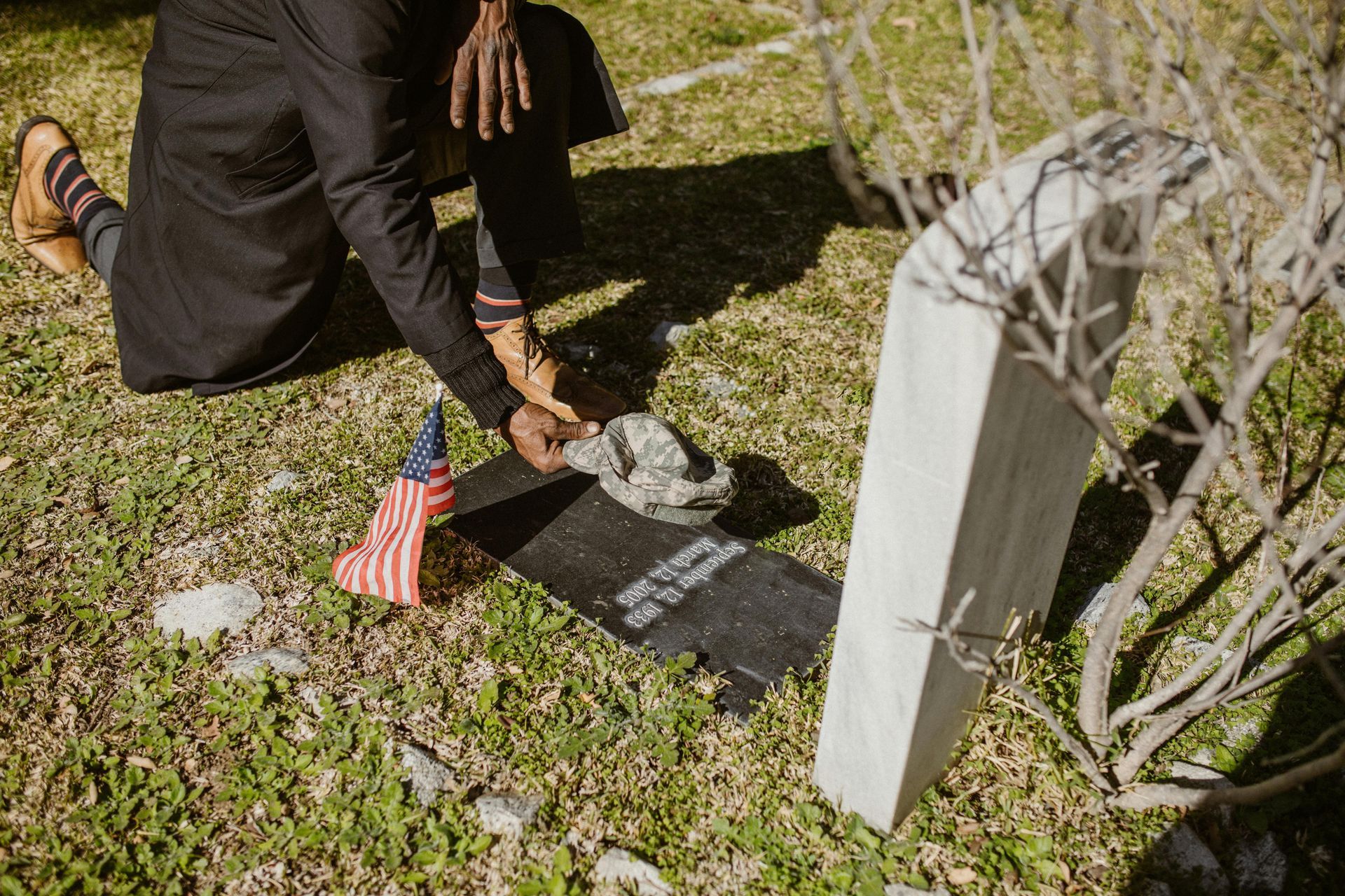Person kneeling at a grave, placing a rock. American flag nearby. Cemetery setting, sunny.