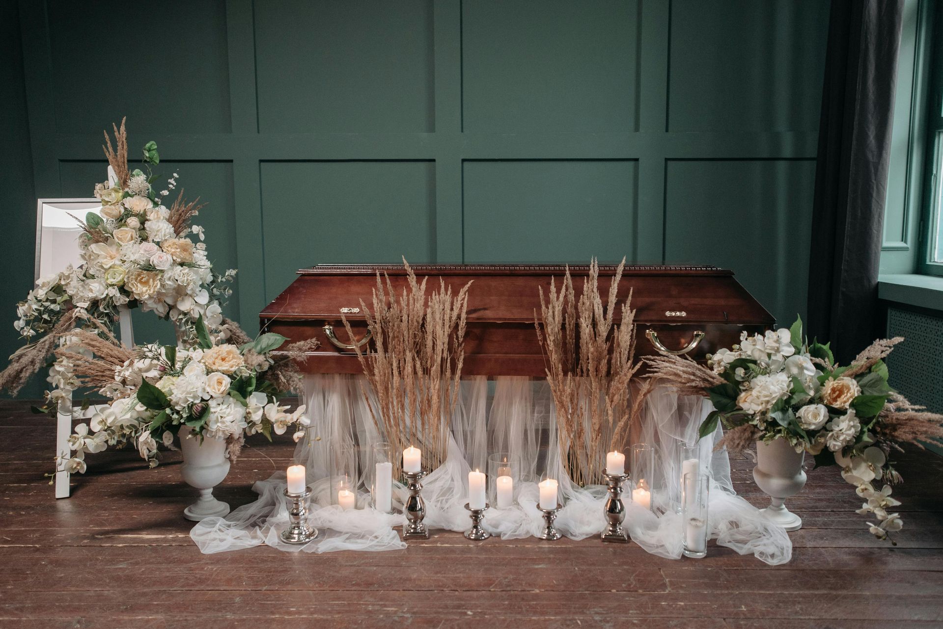 Coffin with floral arrangements, candles, and drapery on a wooden floor in front of a dark green wall.