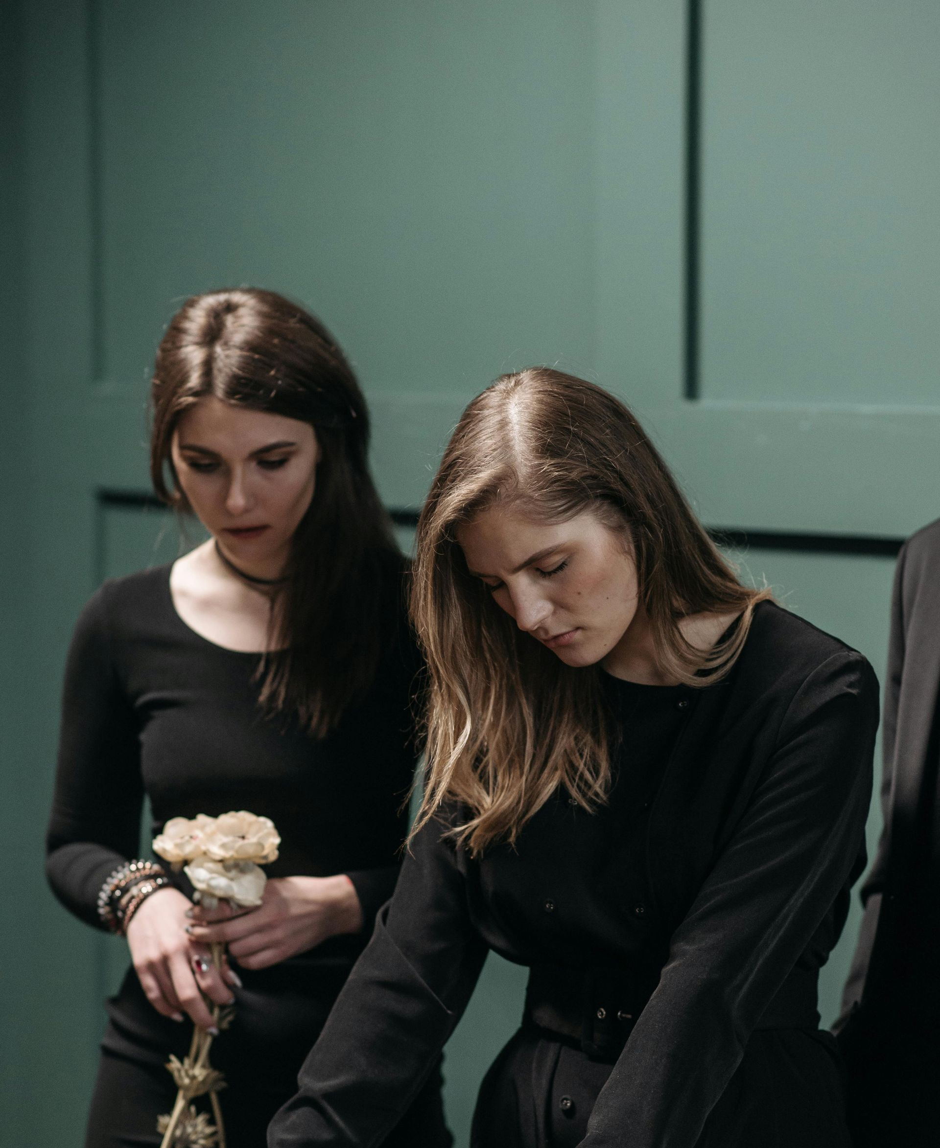 Two women in black, heads bowed, looking down. One holds flowers. Teal wall.