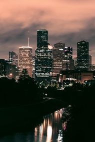 Night cityscape with lit skyscrapers reflecting in a dark river, cloudy sky overhead.