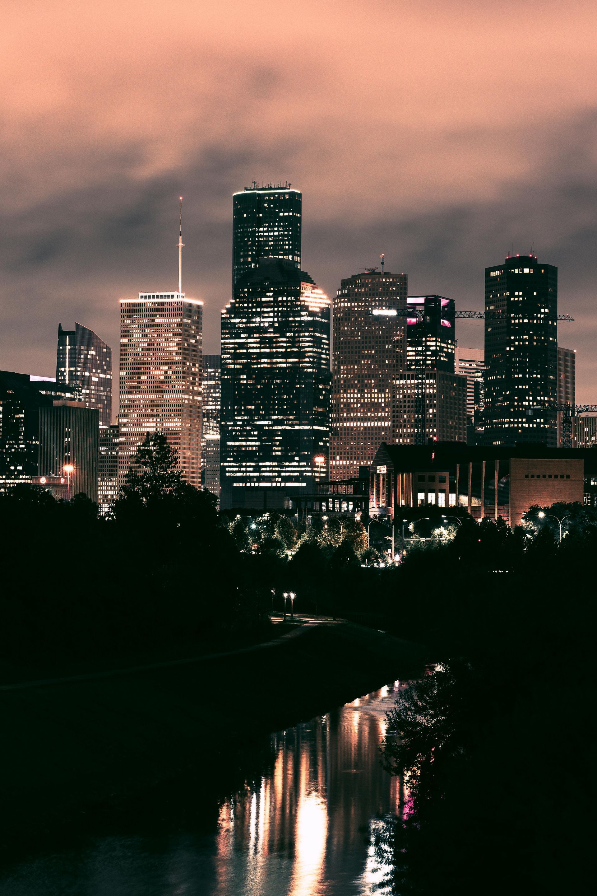 Night cityscape with lit skyscrapers reflecting in a dark river, cloudy sky overhead.