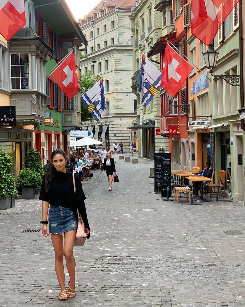 Woman walking down a cobblestone street lined with buildings, Swiss flags, and outdoor seating.