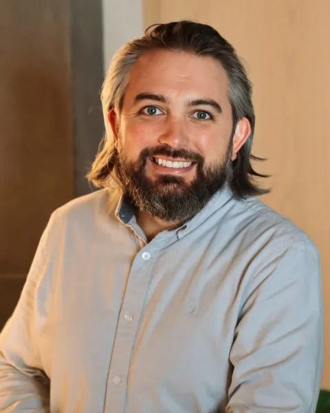 Man with beard smiles, wearing a gray button-down shirt.