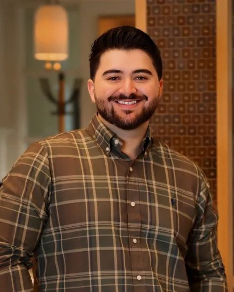Man with beard smiles, wearing plaid shirt, indoors.