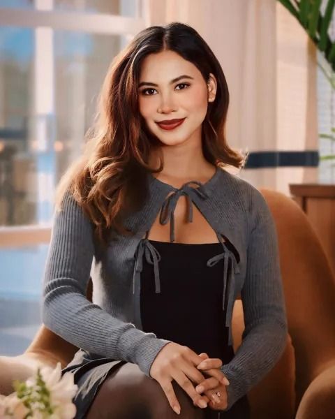 Woman with brown hair, wearing a gray cardigan over a black dress, sitting, and smiling in a room.