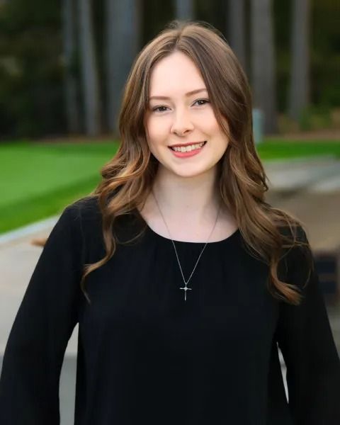 Woman with long brown hair smiling, wearing a black shirt and a silver cross necklace, outdoors.