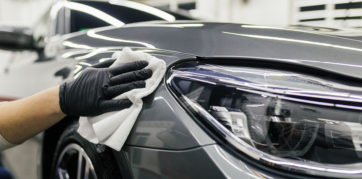 Person in black gloves wiping a gray car's headlight with a white cloth.