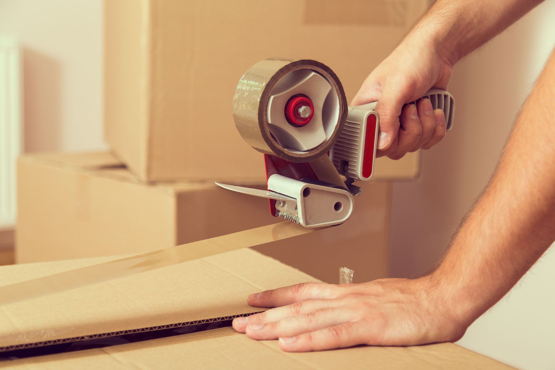Manos sellando una caja de cartón con un dispensador de cinta adhesiva. Cajas apiladas al fondo.