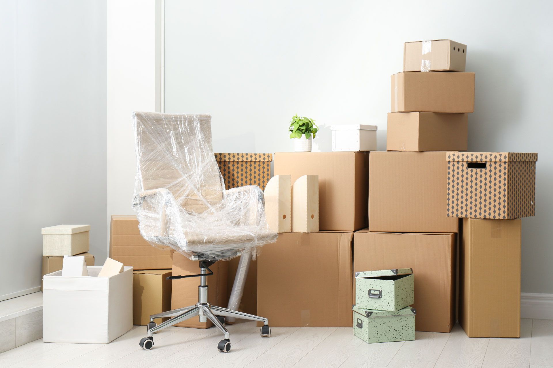 Una silla de oficina envuelta en plástico, rodeada de cajas de cartón, sugiere una mudanza. Una pequeña planta reposa sobre una caja.