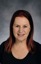 Woman with red-tinted hair smiles at the camera. She wears a black top in front of a gray backdrop.