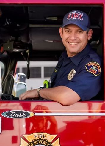 Firefighter in a red fire truck, smiling. Wearing a blue uniform and hat. City of Beverly Hills logo visible.