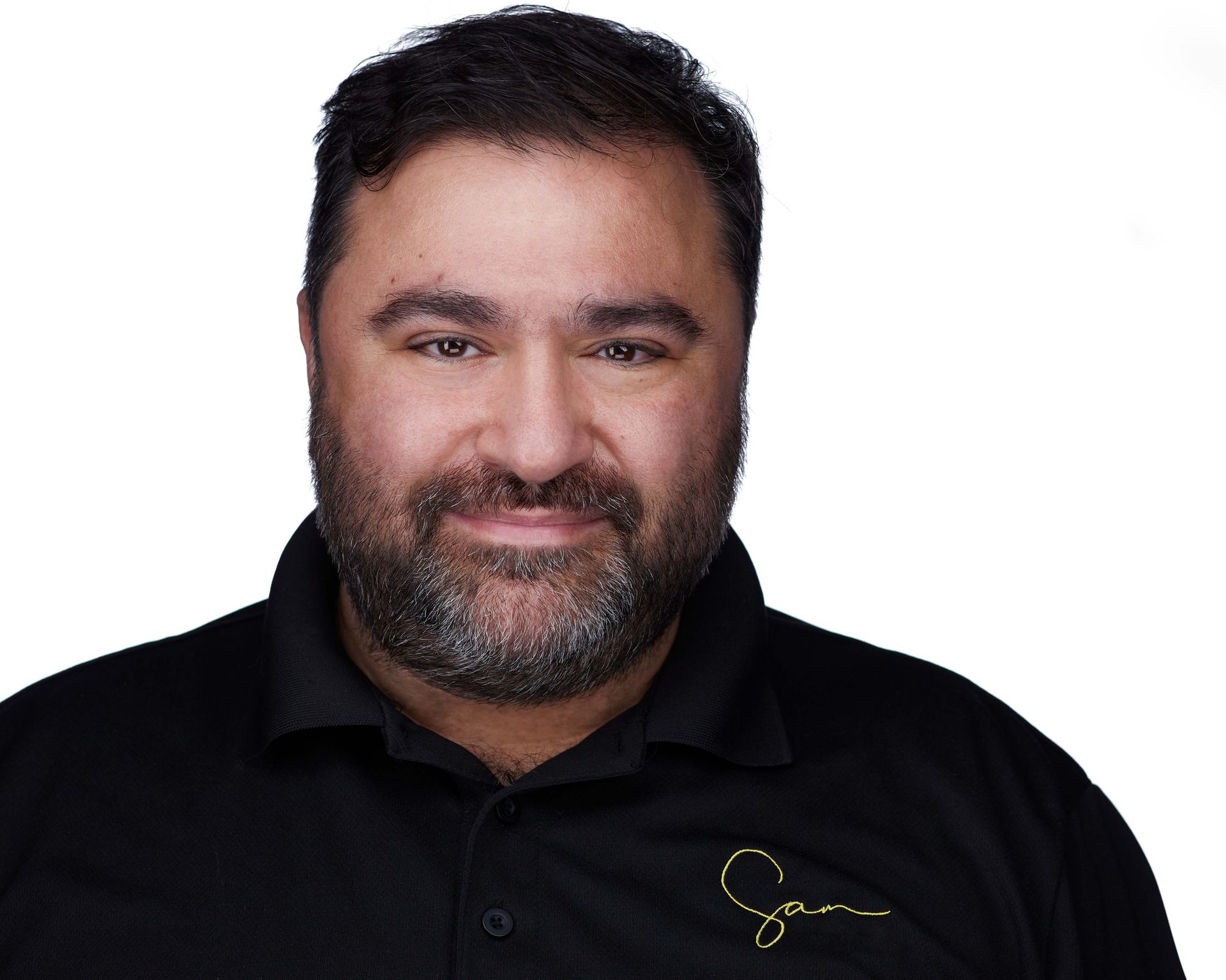 Sam Fatima, Photographer Man with a beard in a black shirt smiles against a white background.
