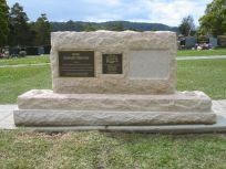 A light-colored stone monument with two plaques and a blank space on it, situated in a grassy, open cemetery setting.
