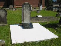 A weathered tombstone with a smaller, modern black plaque sits atop a bright white concrete slab in a grassy cemetery.