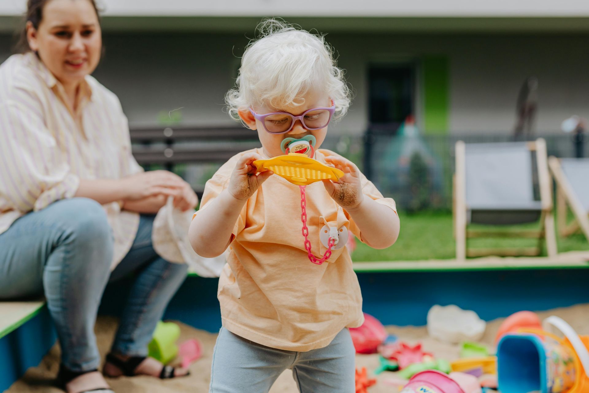 A little girl with glasses is playing in a sandbox while a woman sits on a bench.