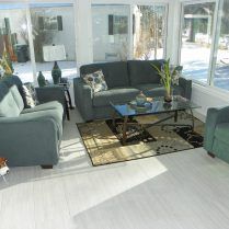 A sunroom with a gray sofa and armchairs, a glass coffee table, and a patterned rug on a light wooden floor.
