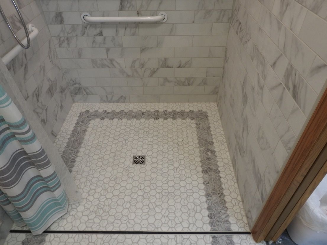 Accessible shower with marble-look tiled walls and hexagon tile floor. Includes grab bar and border.