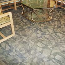Close-up of carpet with swirling gray and tan patterns near a gold and glass coffee table and wooden chair.