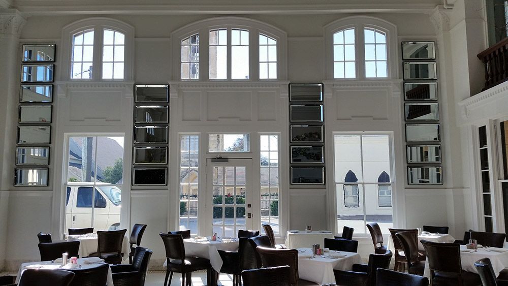 Bright dining room with tall arched windows, white walls, and tables and chairs in front of a sunlit view outside