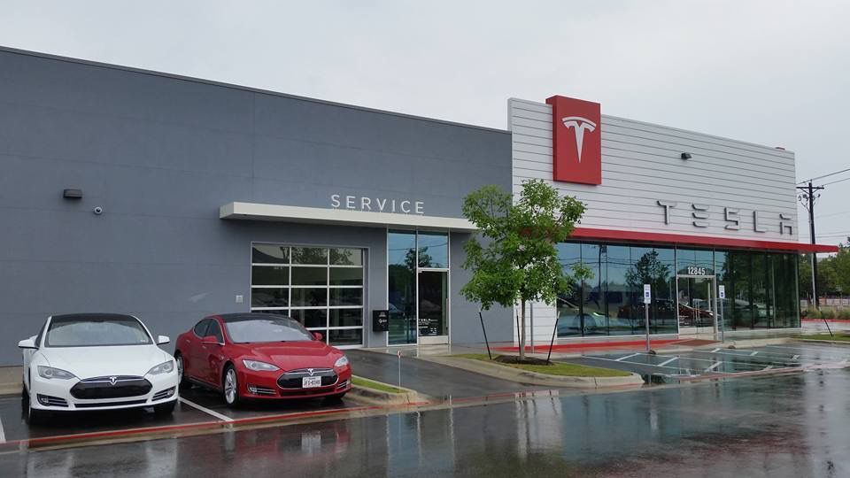 Tesla dealership exterior on a rainy day, with parked cars and a glass storefront.