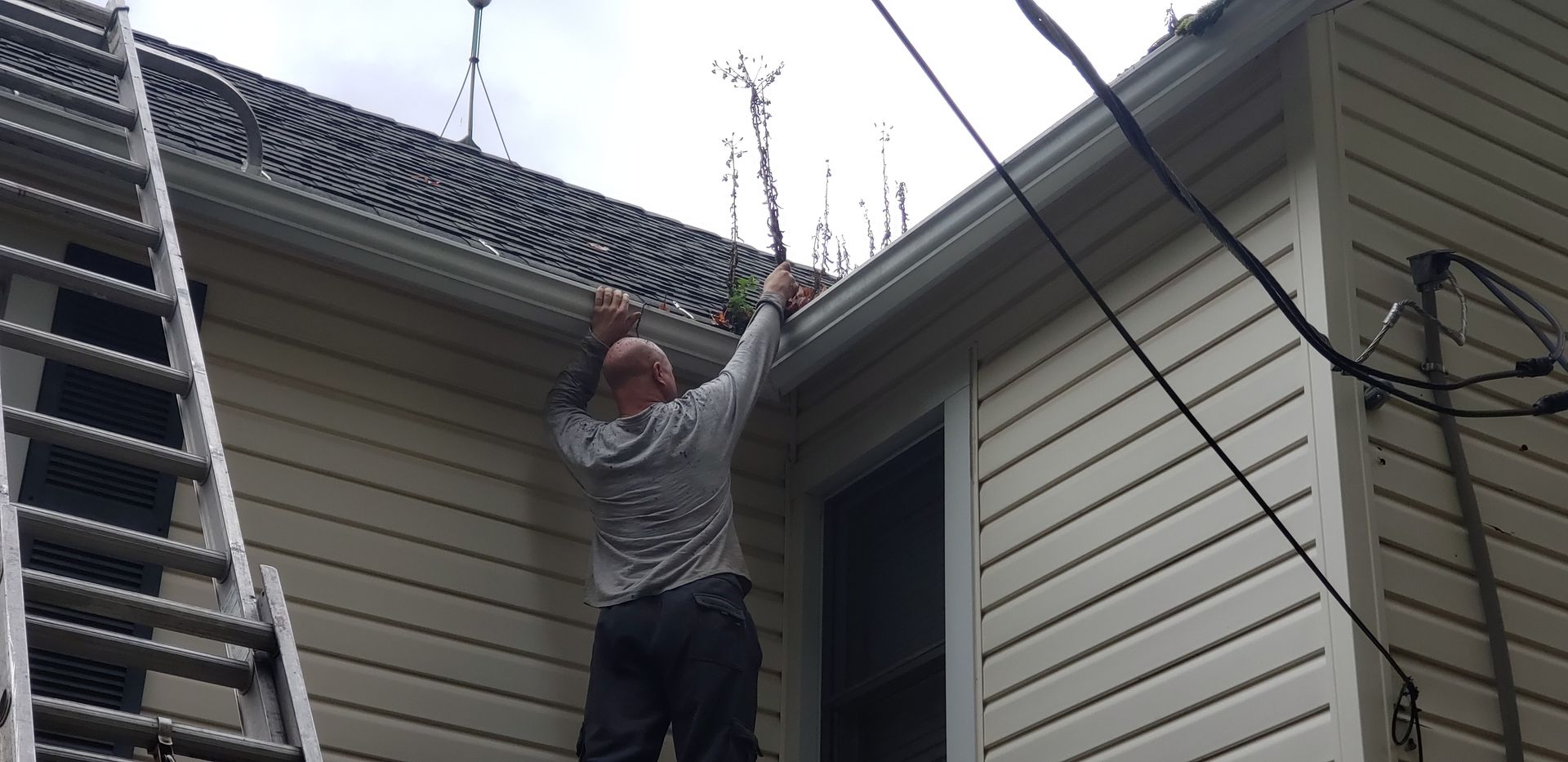 A person stands on a ladder, clearing debris and vegetation from the rain gutters on the side of a tan-sided house.