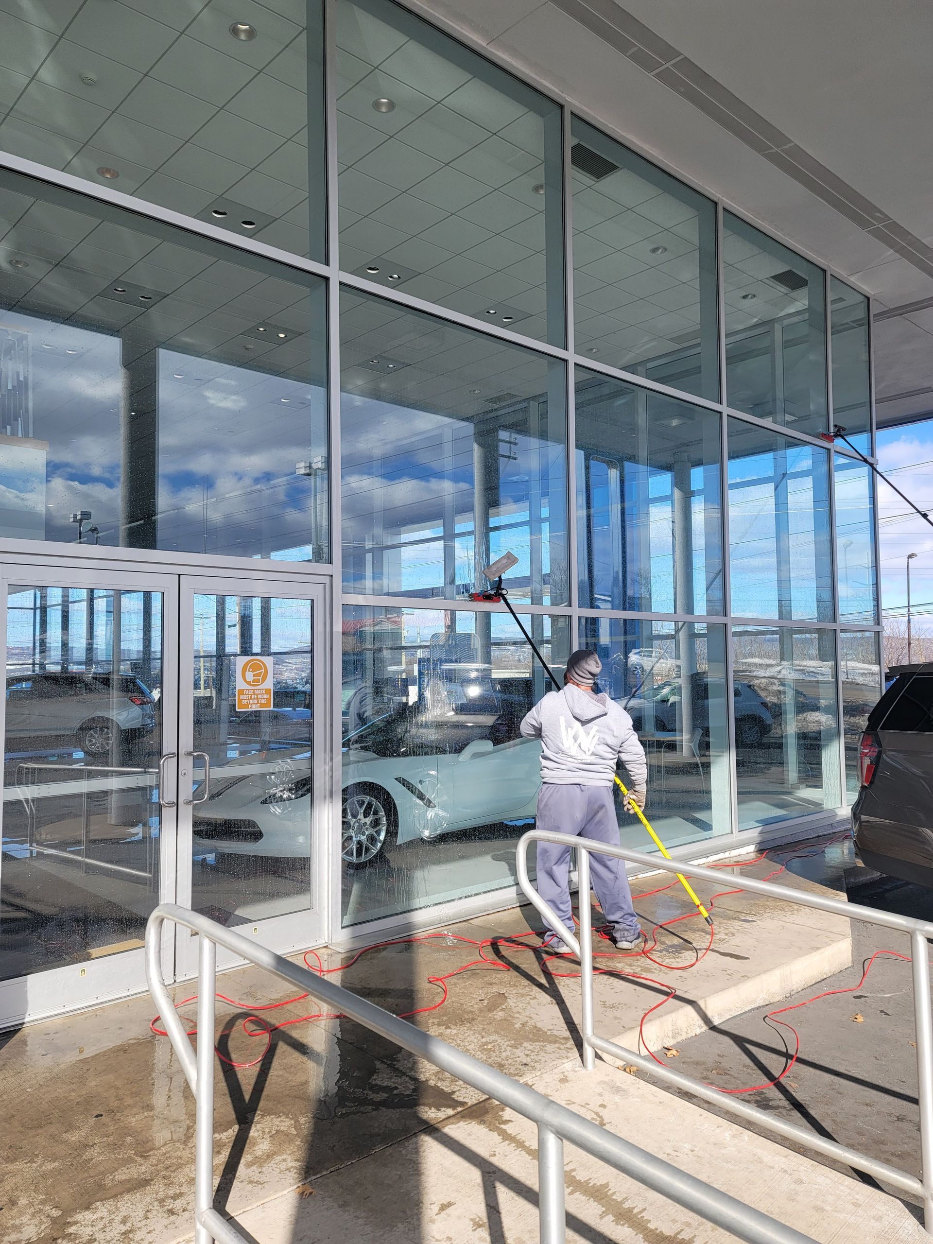 A person in gray clothes uses a long-handled tool to clean the large glass windows of a car dealership.