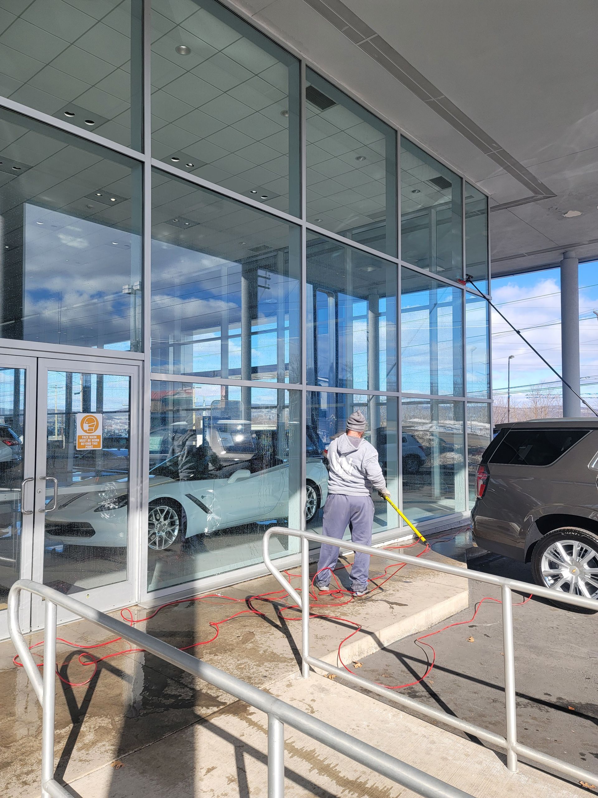 A person in casual clothes uses a long pole to clean the large glass windows of a car dealership on a sunny day.