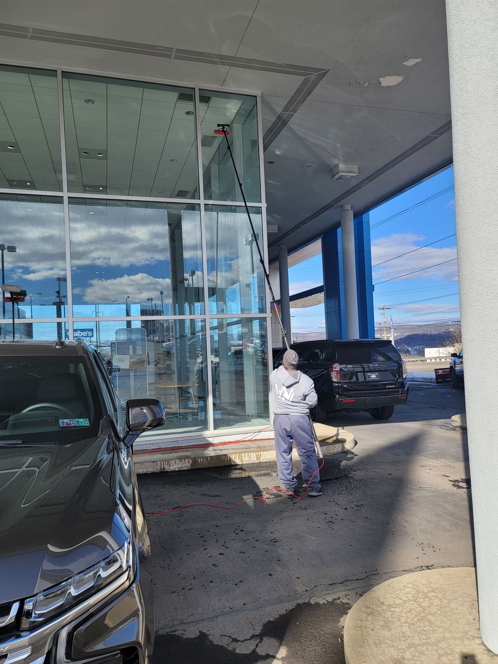 A person cleans the exterior glass of a dealership building using a long-handled brush under a covered parking area.