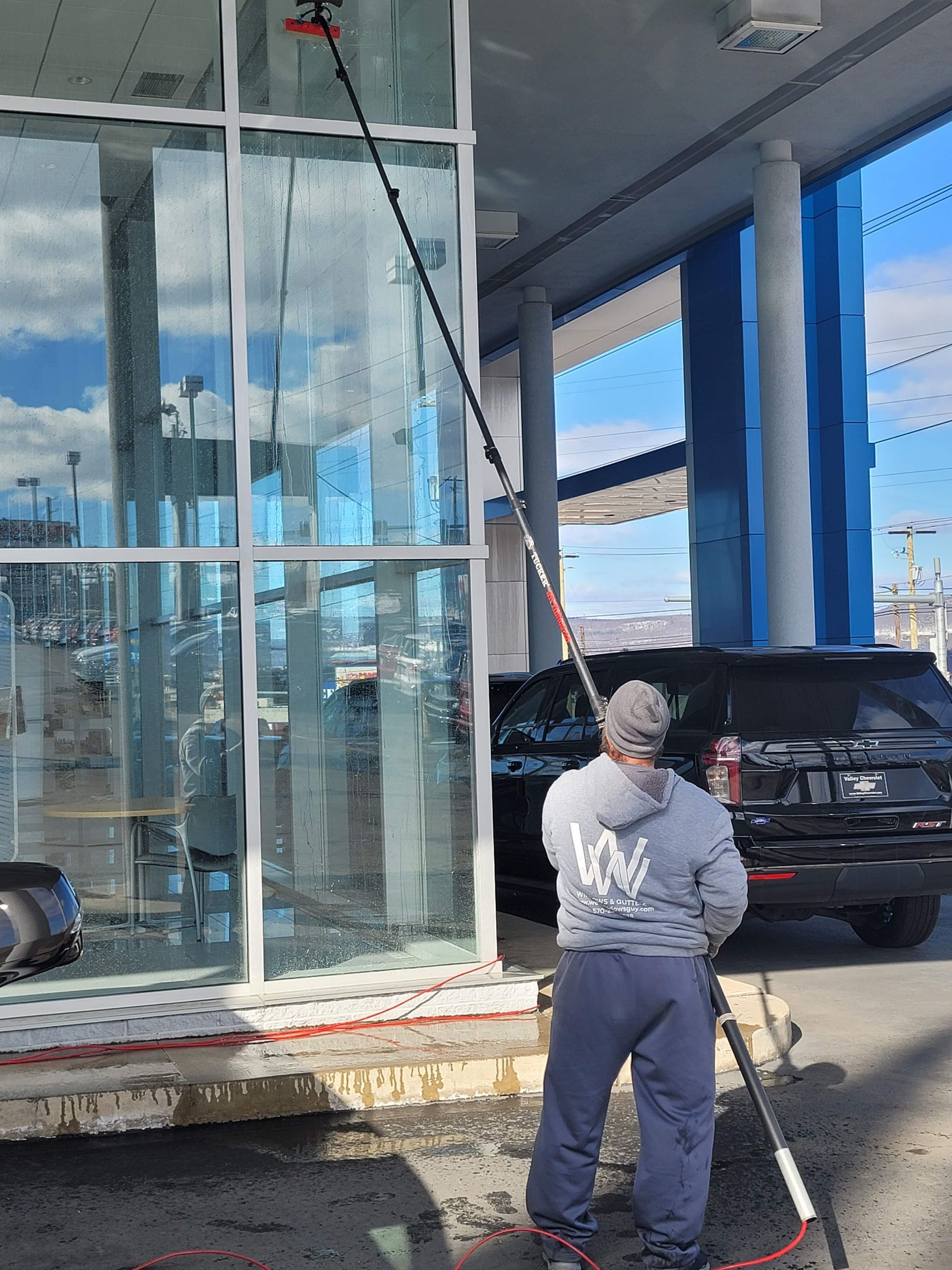 A person in a gray hoodie uses a long-reach pole to clean the exterior windows of a building.