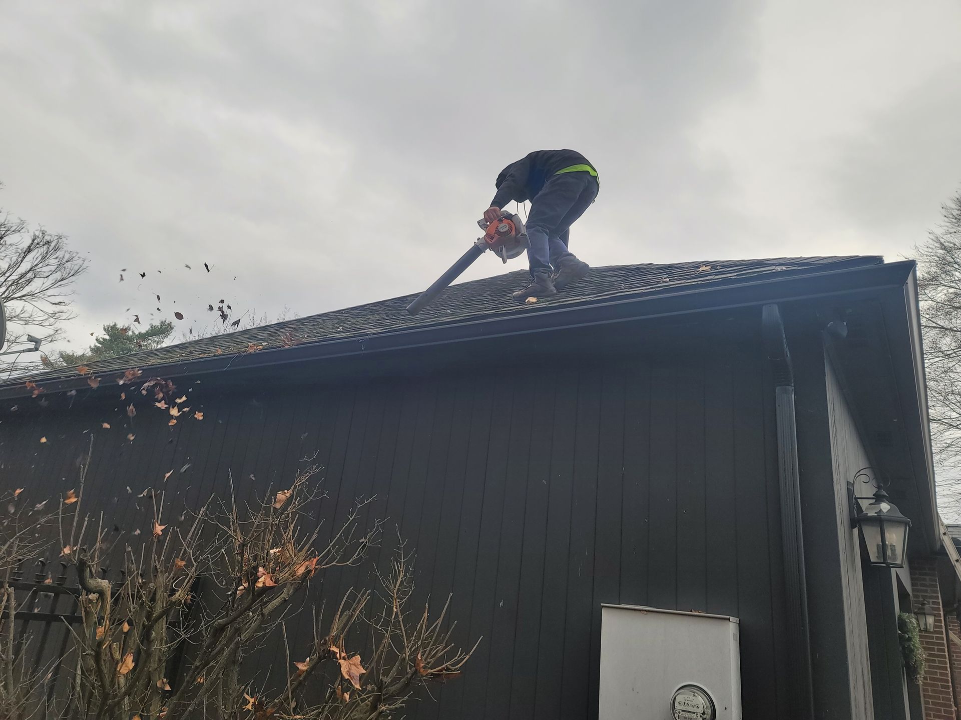 A person stands on the sloped roof of a dark building, using a leaf blower to clear debris from the shingles.