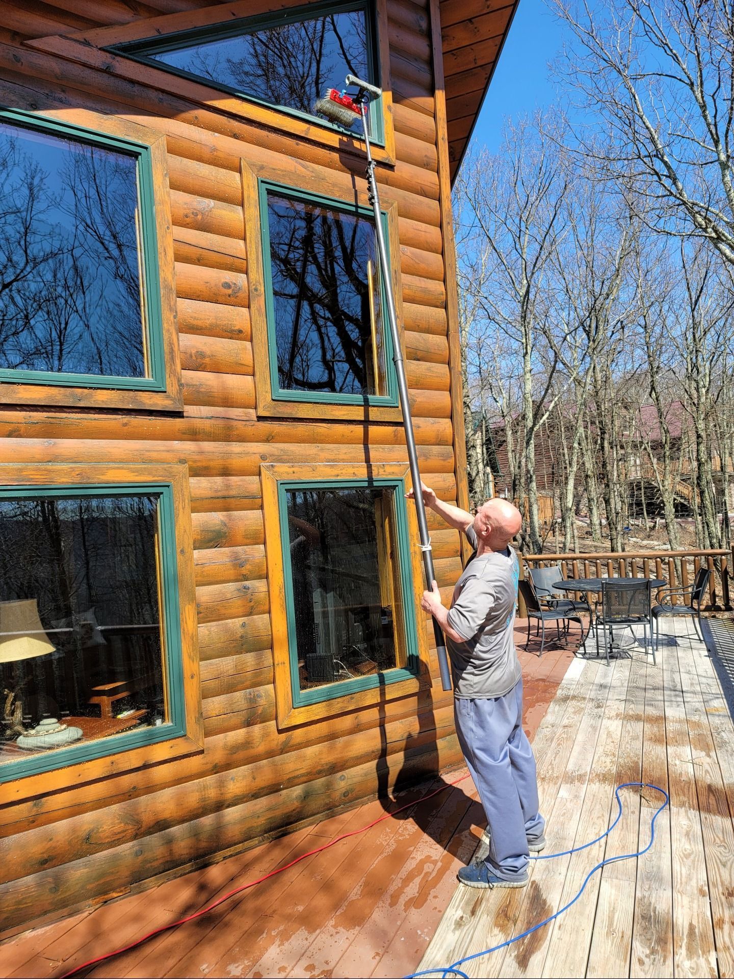 A person uses a long extendable pole with a brush to clean the upper windows of a wooden log cabin on a sunny day.