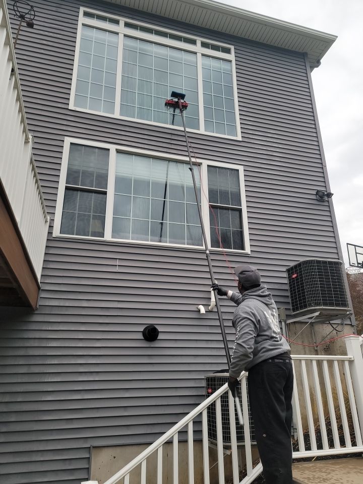 A person stands on a deck using a long pole to clean the exterior second-story windows of a gray-sided house.